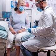 Doctor reading diagnosis of senior woman illness wearing face mask against covid pandemic as safety precaution in hospital. Healthcare medical physician consultation during COVID-19 global crisis.