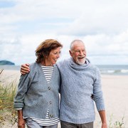 Happy senior woman and man embracing and walking outdoors on sandy beach in autumn.