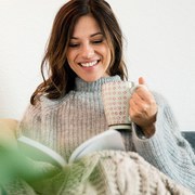 Woman sitting on couch, wrapped in a blanket, reading book, drinking tea